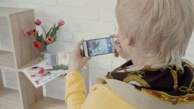 Olgun kadın onun smartphone üzerinde fotoğraf kompozisyon olun. Tasarımcısı içinde beyaz photostudio iş ve rach beyaz tuğla duvarın üzerinde çiçek ve dergi fotoğraf olun. Fotoğraf yapma süreci