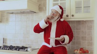 Noel Baba kırmızı kostümlü Mutfak mobilya üzerine oturur. Yaşlı adam beyaz plaka kurabiye yiyor ve cam içecekler süt.