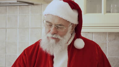 Noel Baba kırmızı kostümlü Mutfak mobilya üzerine oturur. Yaşlı adam kurabiye yiyor ve cam içecekler süt.