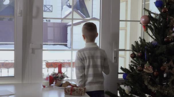 小男孩呆在白色起居室的桌子旁边 看着窗外 看到圣诞老人带着一袋礼物来了 孩子转过身来 脸上带着快乐的微笑看着相机 孩子是非常 — 图库视频影像