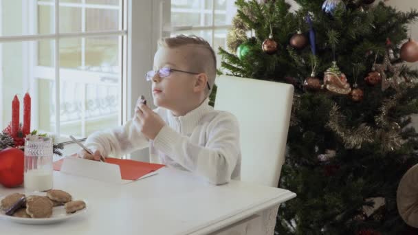 窓際に座って幸せな少年は赤い紙にサンタさんへ手紙を書いて クッキーを食べる 子供は 手書きで焦点を当てて 頭をかきます 子供が笑うと非常に幸せ — ストック動画