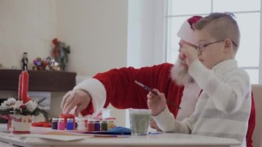 Noel Baba kırmızı kostüm ve soyulmuş ceket ve gözlük küçük çocuk farklı renklerde akrilik boya ile çizin. Şişe mavi boya ve Ihtiyar noktasında çocuk arkadaşı için kapağı açın