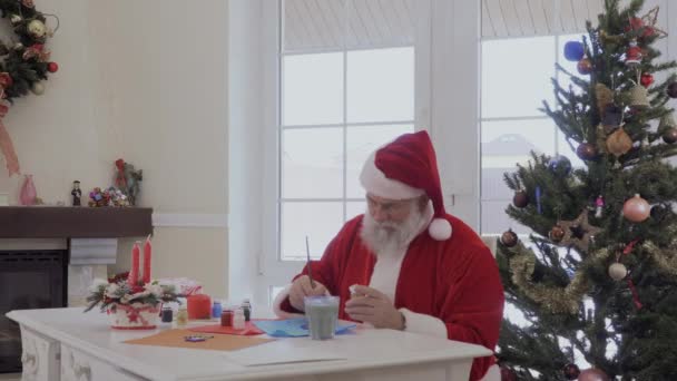 ウィンドウとクリスマス ツリーの近くに座っているサンタ クロースはアクリル絵の具でペイントします 老人ベージュ ペイント入りのボトルを選択し ブラシをかける 水でガラスのブラシをきれい Graybeard を置くクリーン ブラシ — ストック動画