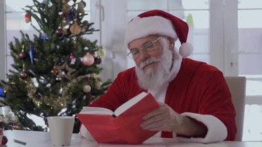 Noel Baba Noel şapka ve masada oturan kırmızı kostüm. Dedesi beyaz bir bardaktan içiyor ve bir kitap okuma. Noel ağacı civarı olduğunu.