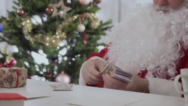 白い長いひげのサンタ クロースがテーブルとクリスマス ツリーの近くカウントお金の上に座る — ストック動画