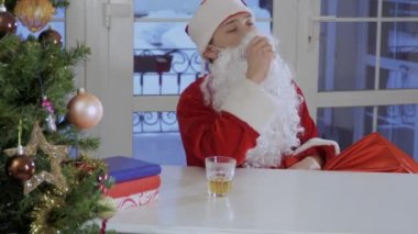 Takma sakal ile Noel Baba kıyafetli sıkılmış adam oturup tablo ve güçlü alkol bir kadeh içmek. Üzgün ve yorgun erkek.
