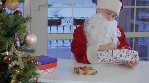 穿着圣诞老人红色服装的年轻人坐在桌子上 把签名放在礼物的一个上面 人握手礼品盒 — 图库视频影像