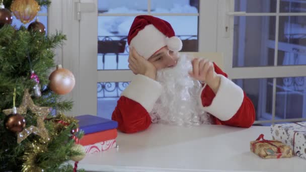 穿着圣诞老人服装的年轻人坐在桌子旁边 和绒球的圣诞老人红帽子玩耍 这个人很无聊 — 图库视频影像