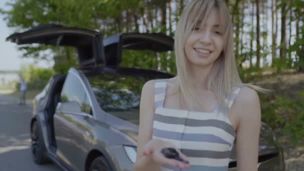 Mladá usměvavá dívka s klíčky na dlaň. Moderní automobil s otevřením dveří stánku rozostřeného pozadí. Zpomalený pohyb.