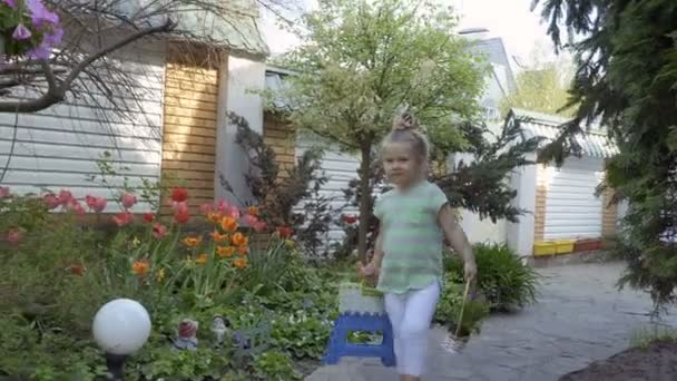 Little girl walking with basket of flowers, toy and chair on path near house — Stock Video