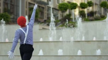 Kırmızı bere içinde komik MIME Çeşme bakcground ve ellerini bir iletken hareketli bir performans vardır. Sökülen forması giyen sokak aktör ve oyun komik sahneler insanlar için be.