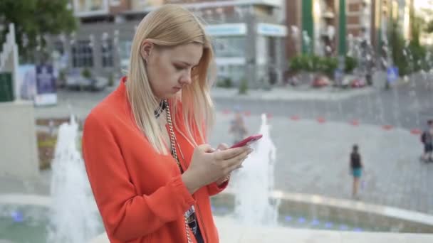 Ganska snygg ung flicka i röd jacka med hennes telefon vid fontänen bakgrund — Stockvideo