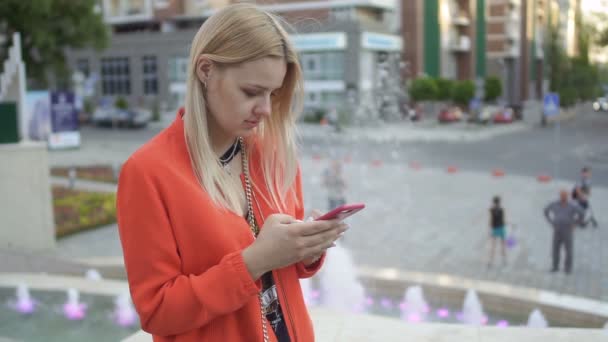 若いかわいらしい噴水の背景で彼女の電話の立っている赤いジャケットの女の子 — ストック動画