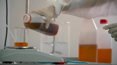 Elektronik ölçekte bir şişesi içine portakal sıvı koyu sıvı dökme kimyager. Deney Laboratuvar donatımı hazırlama ve iki sıvıları karıştırma bilim adamı. İlaç
