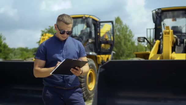 Менеджер в солнцезащитных очках и с папкой в руках стоят на заднем плане тракторов — стоковое видео