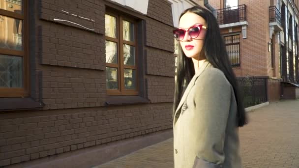 Стильная молодая женщина в солнцезащитных очках и пальто на фоне коричневого здания на улице — стоковое видео