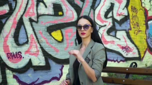 Femme élégante en lunettes de soleil assise sur un banc contre un mur avec des graffitis de rue — Video