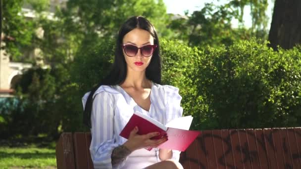 O femeie frumoasă în ochelari de soare citește cartea în parc — Videoclip de stoc