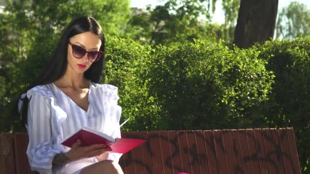 Bruna elegante in occhiali da sole leggere il libro su una panchina nel parco — Video Stock