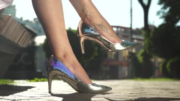 Žena v elegantní stříbrné boty na vysokých podpatcích sedí na lavičce v parku — Stock video