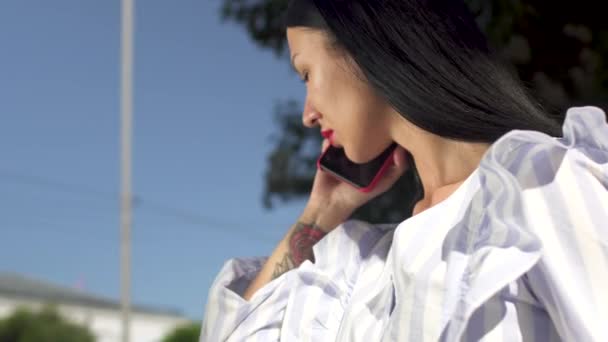 Elegante morena con lápiz labial rojo hablando por teléfono contra el cielo — Vídeo de stock