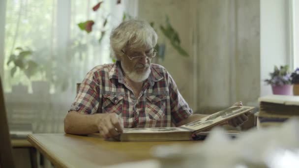 Старший мужчина сидит дома и смотрит свой старый альбом с фотографиями — стоковое видео