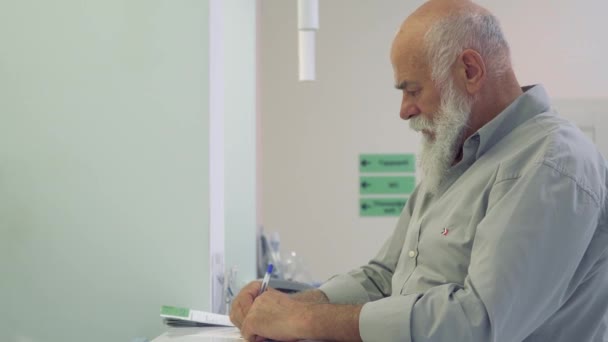 Hombre mayor fiil el formulario en la recepción en el hospital y mirando a la cámara — Vídeo de stock