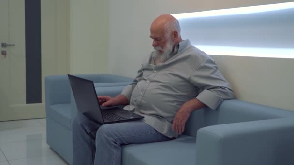 Volwassen man met laptop tijdens het wachten op zijn beurt in kliniek zittend op de Bank — Stockvideo