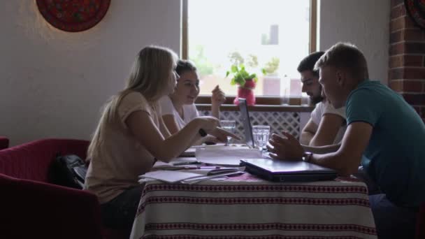 つの若者のグループがテーブル近くのレストランで座って会話を持っている テーブルの上に練習帳 ノート パソコン 一緒に大学で試験の準備を学生 — ストック動画