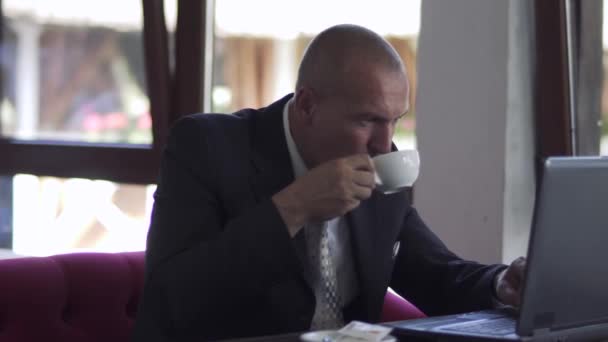 Empresario bebe café sentado en la mesa en el restaurante y trabajando en el portátil — Vídeos de Stock