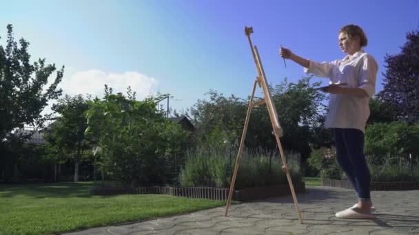 Blonde girl paints on an easel outdoors — Stock Video