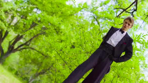 Όμορφος άνθρωπος στο κοστούμι βόλτες στα ξυλοπόδαρα στο πάρκο κοντά στο δέντρο — Αρχείο Βίντεο