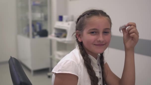 Девочка-подросток держит слуховой аппарат в руке и смотрит в камеру — стоковое видео