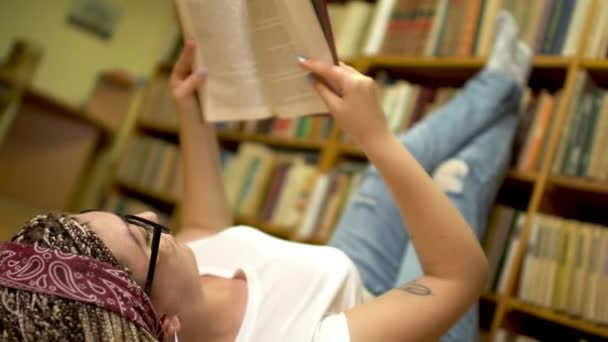 Hermosa chica está leyendo un libro tirado en el suelo en la biblioteca . — Vídeos de Stock