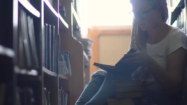 Young girl is sitting on the floor between rows of books — Stock Video