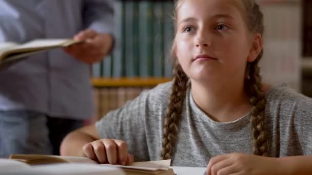 Doce menina está sentada a uma mesa na biblioteca — Vídeo de Stock