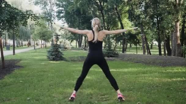 在公园做早操的漂亮女孩 小女孩跳起来 把她的胳膊和腿放到一边 漂亮的年轻金发女郎在运动紧身裤和运动 T恤做运动 — 图库视频影像