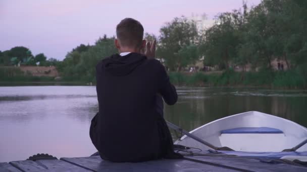 Un tipo guapo se sienta en un muelle cerca de un barco — Vídeos de Stock