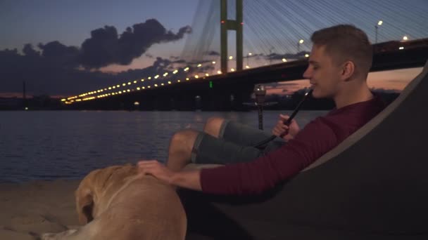 Beau mec assis avec un chien sur la plage et fume un narguilé — Video