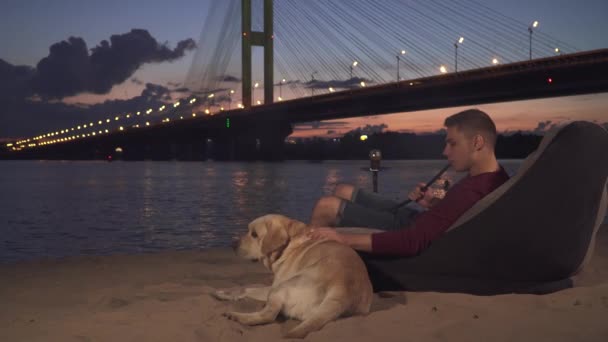 Jonge kerel zit met een hond op het strand en rookt een waterpijp — Stockvideo