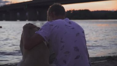 Genç adam bir köpekle nehir kıyısında oturuyor