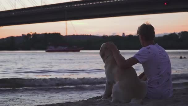 El joven está sentado con un perro en la orilla del río. — Vídeo de stock