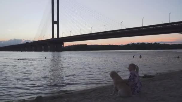 Menina bonita está sentada com um cão na margem do rio — Vídeo de Stock