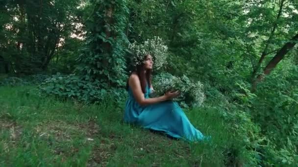 Красива дівчина в довгій сукні з вінком сидить у лісі — стокове відео