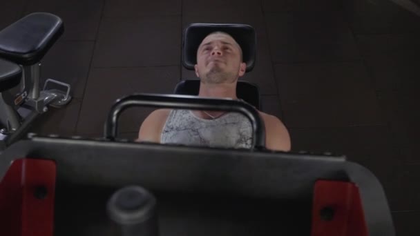 健身房里的帅哥穿着 T恤衫和短裤的运动男正在一个训练大厅里 那家伙在模拟器上躺在他的背上 年轻人正在用模拟器做腿部练习 视图从 — 图库视频影像
