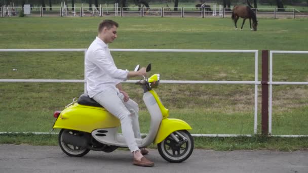 若い男は原付けに座っているし 馬に見えます ハンサムな男はスクーターに座っています 黄色のレトロなモーター自転車の若い男 — ストック動画