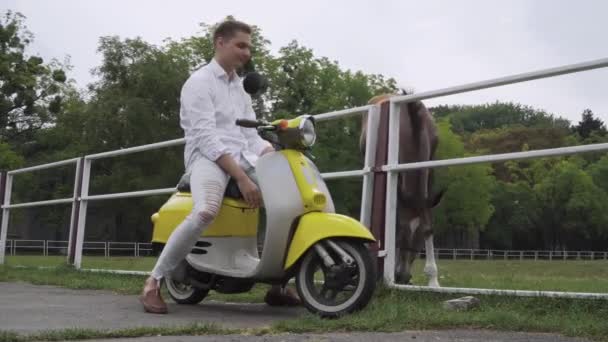 若い男はスクーターに座っているし 馬をストロークします バイクに白シャツでハンサムな男は 黄色のレトロなモーター自転車の若い男 — ストック動画