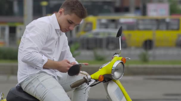 那家伙正在坐在摩托车上的电话里聊天 穿着黄色复古轻便摩托车的白色衬衫的帅哥骑滑板车 — 图库视频影像