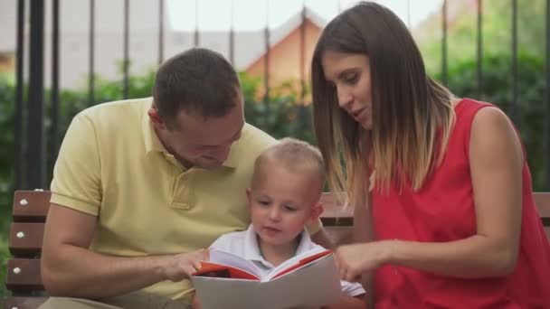Papá, mamá e hijo están leyendo un libro en un banco del parque. — Vídeos de Stock
