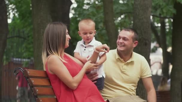 Los padres jóvenes se sientan con un hijo bebé en un banco del parque — Vídeos de Stock